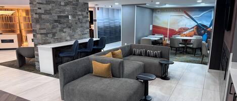 Un moderno vestíbulo de hotel con un sofá seccional gris, una pared de acento de piedra y un área de comedor con mesas y sillas.
