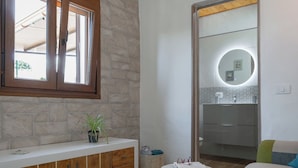 Un baño con pared de piedra, tocador de madera, espejo redondo y una pequeña mesa redonda con un libro y gafas.