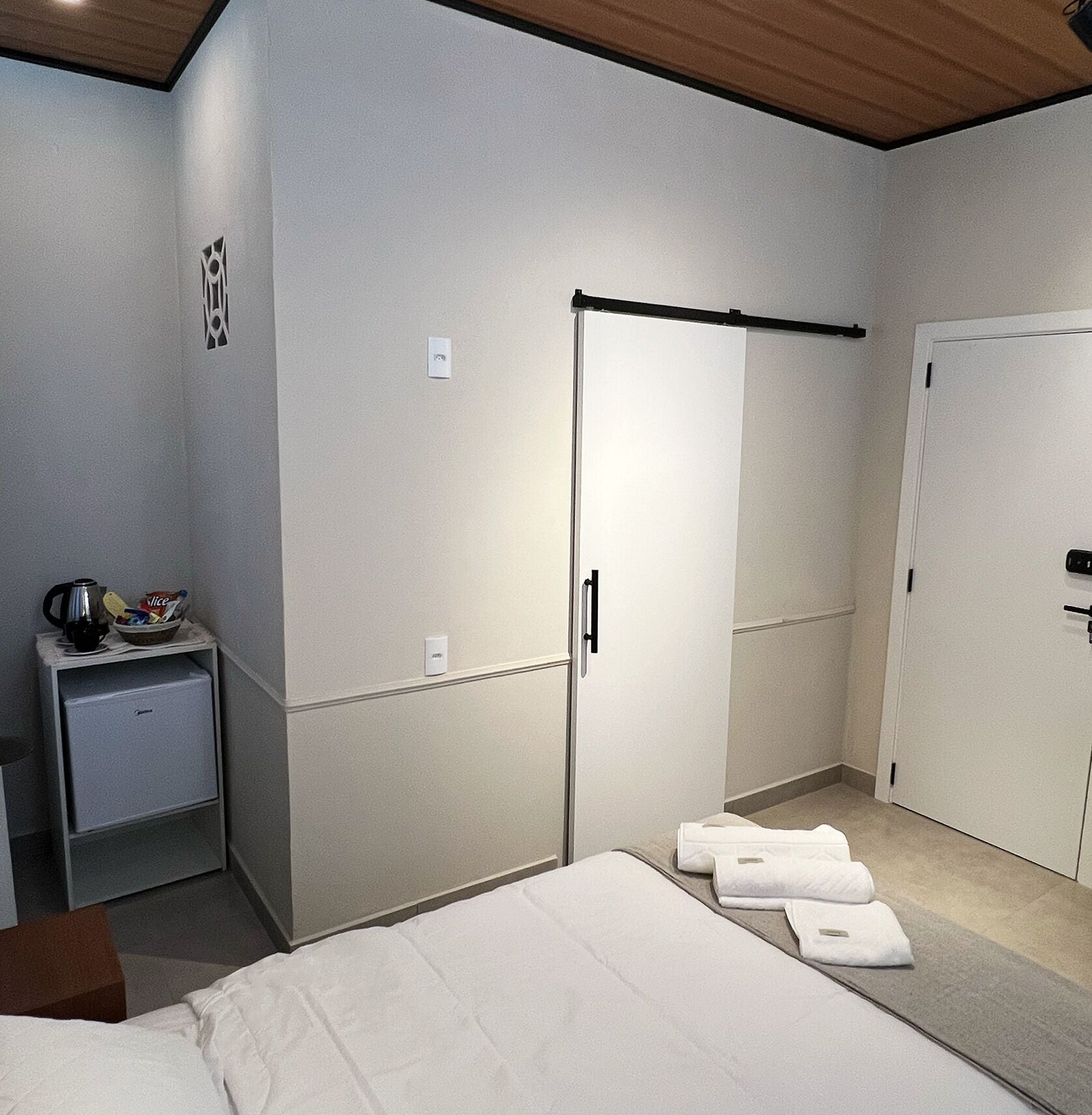 A bedroom with a bed, a wardrobe, a small bedside table with a kettle, and a wall-mounted light.