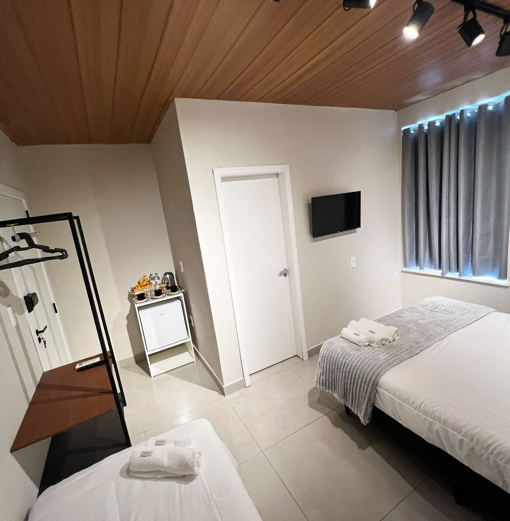 Une chambre d’hôtel avec deux lits, un téléviseur fixé au mur, une petite table avec une bouilloire et des tasses, un porte-manteau et une porte.