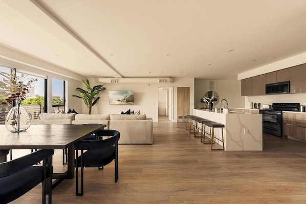 A modern kitchen with a central island, a dining area with a table and chairs, and a living area with sofas and a large window.