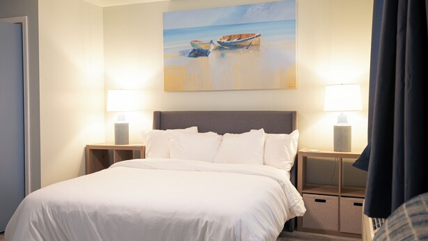 A hotel room with a large bed, two bedside lamps, a painting of a boat on a beach, and a view of the sea. - Anchor Point at Oak Shores! 5-min walk to beach (Biloxi)