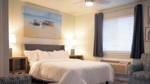 A neatly arranged hotel room with a large bed, two bedside tables with lamps, a wall-mounted air conditioner, a window with curtains, and a painting above the bed. - Anchor Point at Oak Shores! 5-min walk to beach (Biloxi)