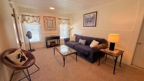 Un salon chaleureux avec un canapé, une table basse en verre, une table d’appoint avec une lampe, un fauteuil agrémenté d’un coussin décoratif, une cheminée et un tableau encadré au mur.