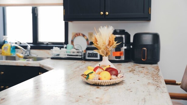 Keittiössä on marmorinen työtaso, hedelmäkori, kuivakukkia sisältävä maljakko, kahvinkeitin ja vedenkeitin.