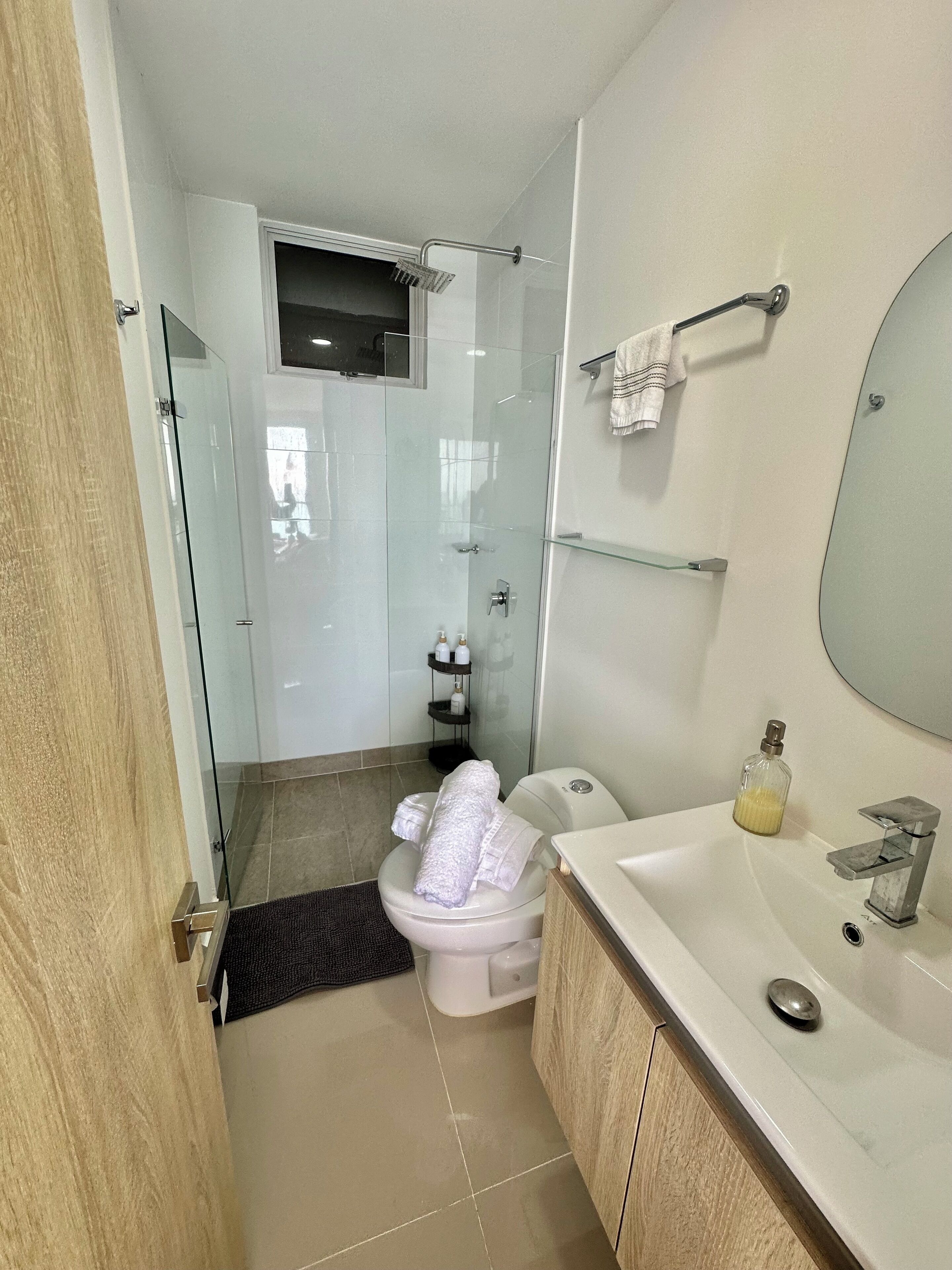 Un baño moderno con ducha de vidrio, inodoro, lavabo con espejo y un pequeño estante con artículos de aseo.