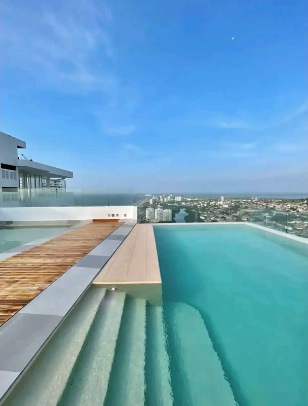 A modern infinity pool with a wooden deck, overlooking a cityscape and the ocean.