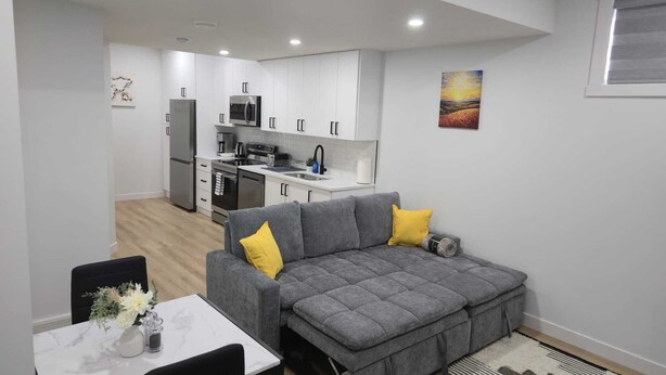 A modern apartment with a grey sofa, a dining table with a vase of flowers, a kitchen with white cabinets, and a painting on the wall. - Mayfair Suite (Airdrie)