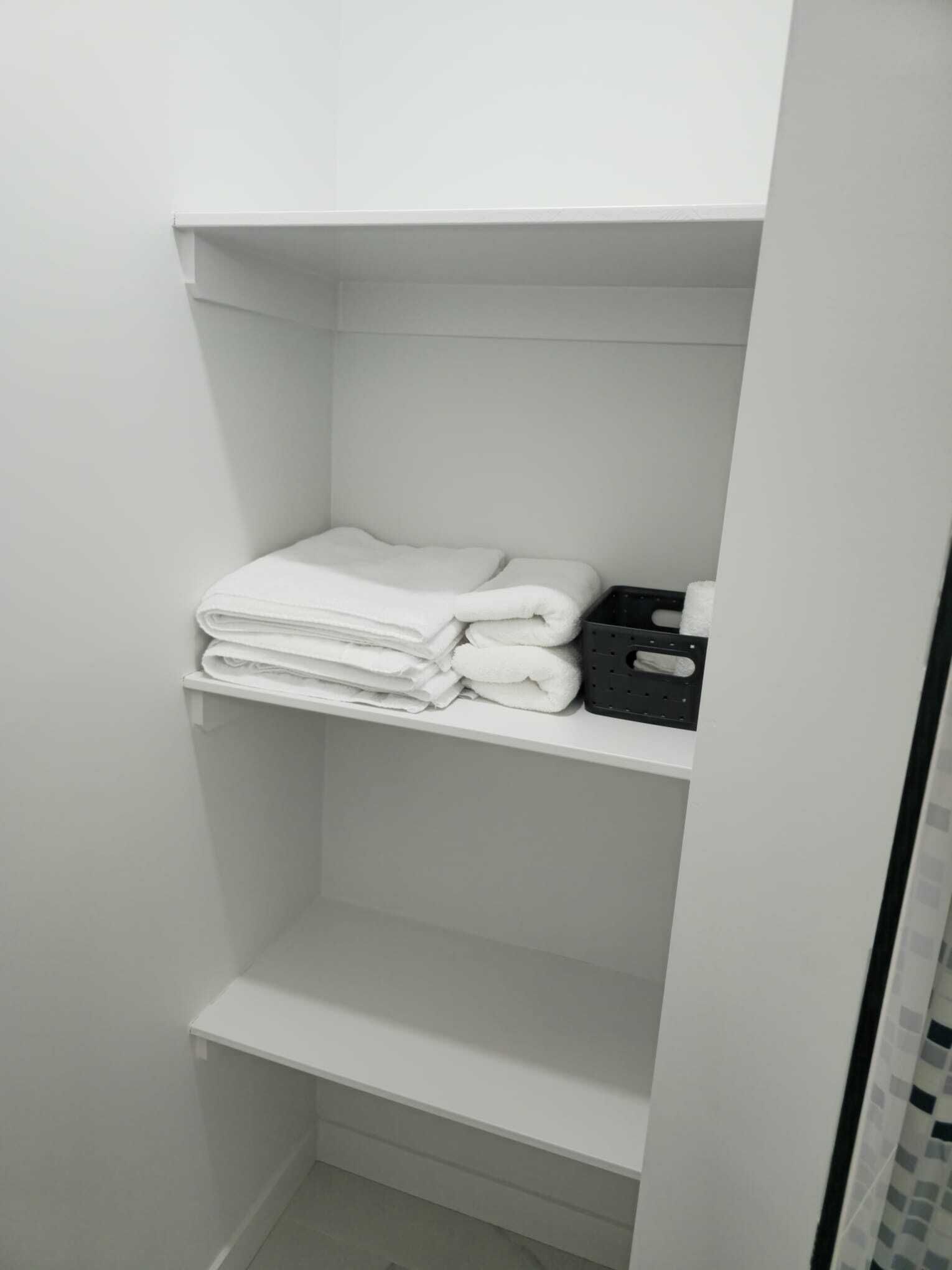 Une salle de bain blanche avec une étagère où sont pliées des serviettes et un plateau noir.
