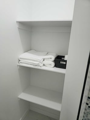 A white bathroom with a shelf holding folded towels and a black tray. - Mayfair Suite (Airdrie)