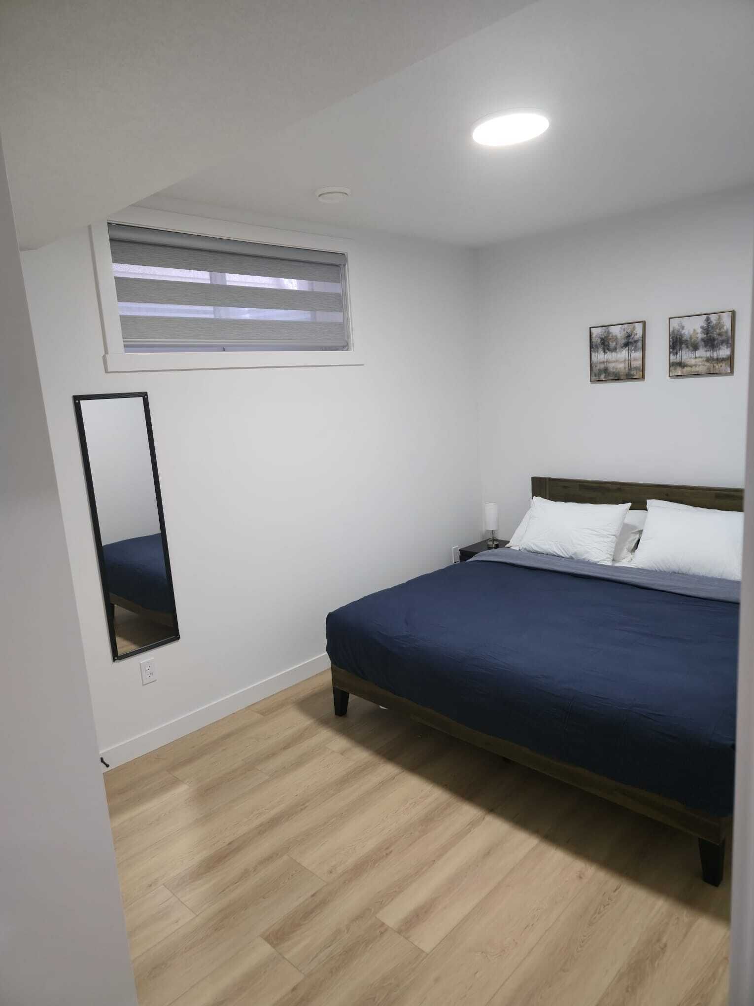 A bedroom with a bed, a window with blinds, a wall-mounted mirror, and two framed pictures.