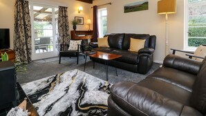 A living room with a leather sofa, a coffee table, a TV, and a view of the outdoors through a sliding glass door. - Barton Lodge (Minehead)