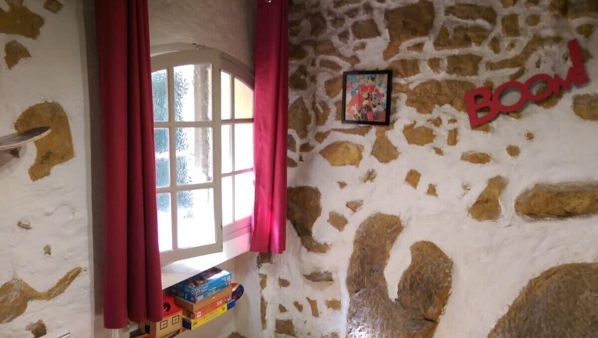 Una stanza con una parete in pietra, una finestra con tende rosa e una mensola piena di libri.