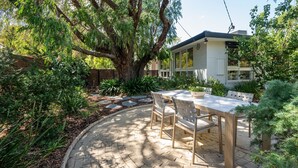 Un patio con tavolo e sedie, circondato da un giardino con diverse piante e un albero grande.