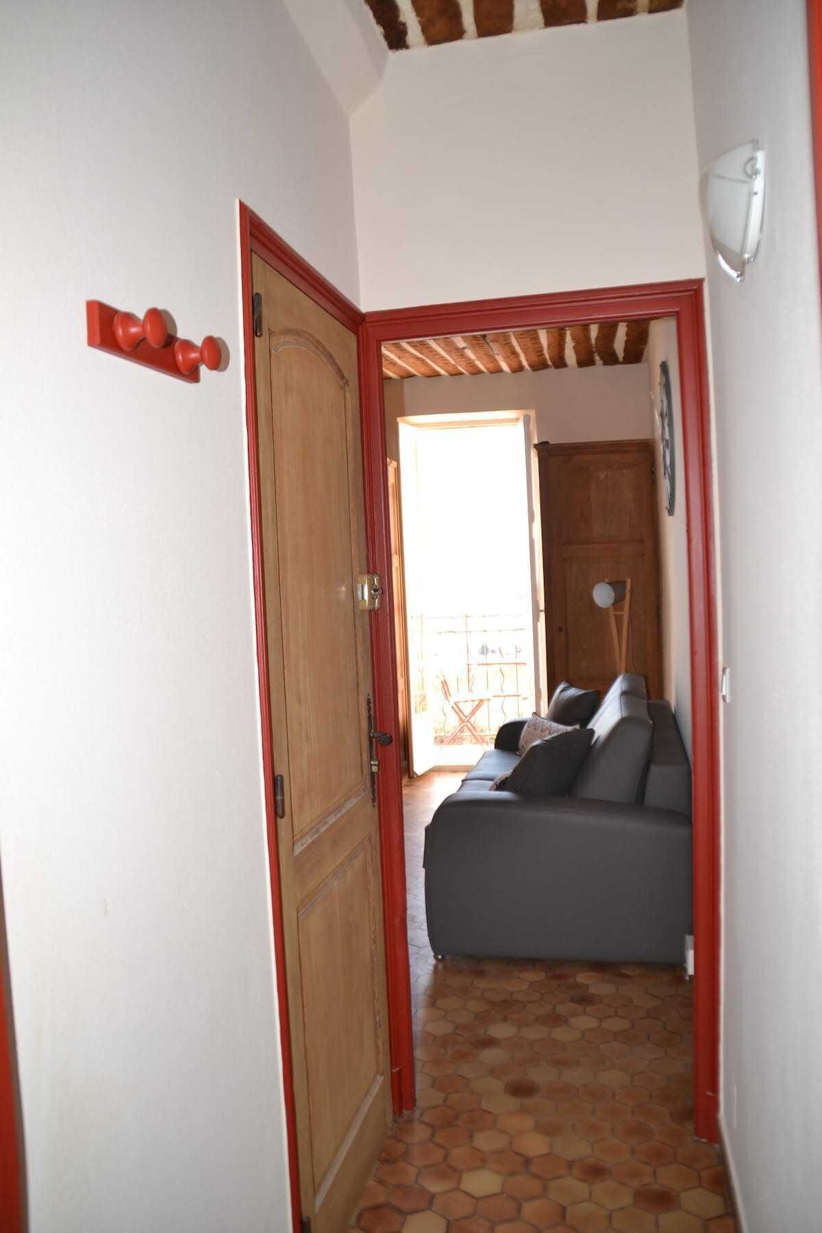 A narrow hallway with a wooden door and a red frame, leading to a room with a gray sofa and a view of the outside.