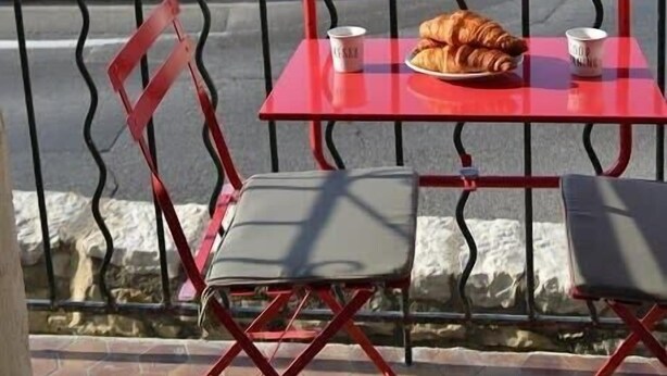 Una mesa roja con dos sillas, un croissant y dos tazas en un balcón con vista a la costa y edificios al fondo.