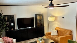 A living room with a television, dark wooden entertainment unit, a green sofa, a brown leather sofa, a white armchair, and a floor lamp.