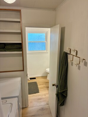 A bathroom with a wooden floor, a white toilet, a window with a view of trees, and a towel rack with a green towel. - Newly Renovated 3BR : Quartz Kitchen : Fast Fiber WiFi : Carport Parking (Campbell)