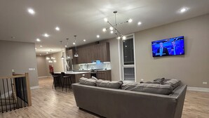 Ein modernes Wohnzimmer mit einer großen, grauen Couch, Holzböden, deckenmontierten Leuchten, einer Küchenzeile mit Barhocker und einem an der Wand befestigten Flachbildfernseher.