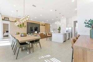 A modern kitchen with a central island, pendant lighting, and a dining area with a long table and chairs. - Paradiso's Luxurious New Home w 15 Ensuites (Orlando)