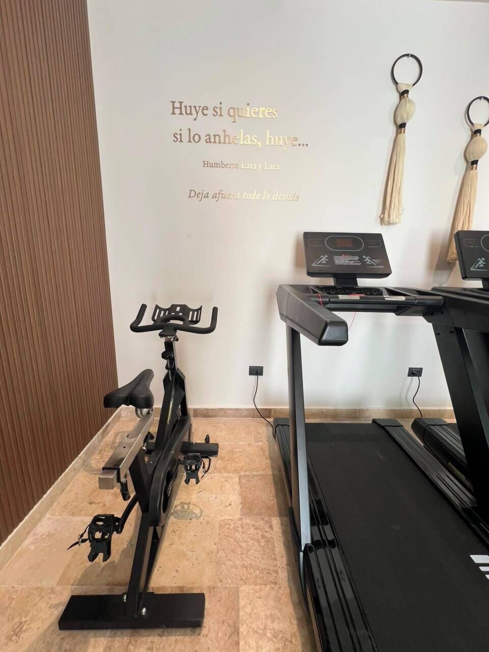 Un gimnasio con bicicletas estáticas y cintas de correr, una frase motivacional en la pared y una puerta de madera.