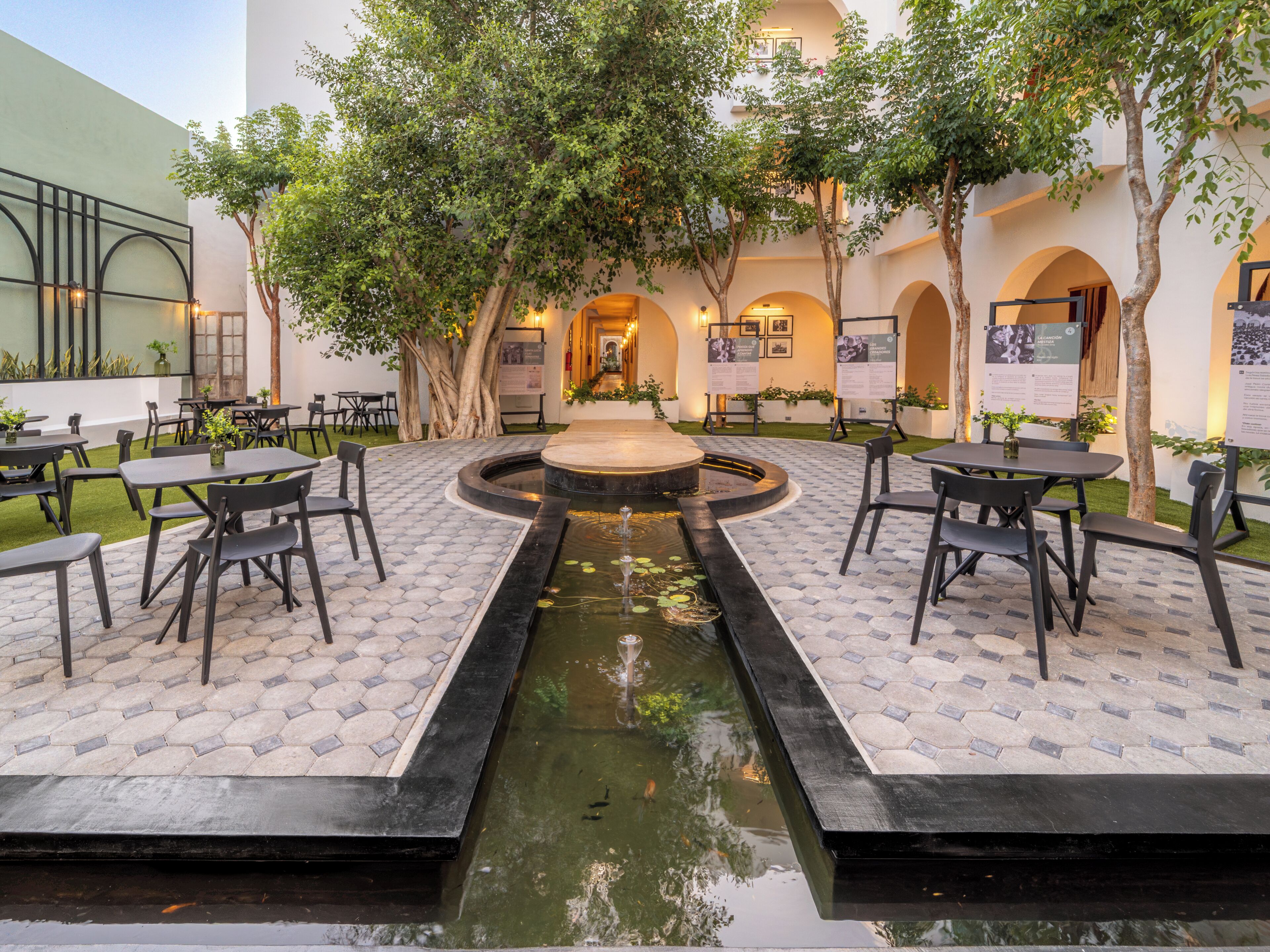 An outdoor patio area with tables and chairs, a central water feature, and trees.