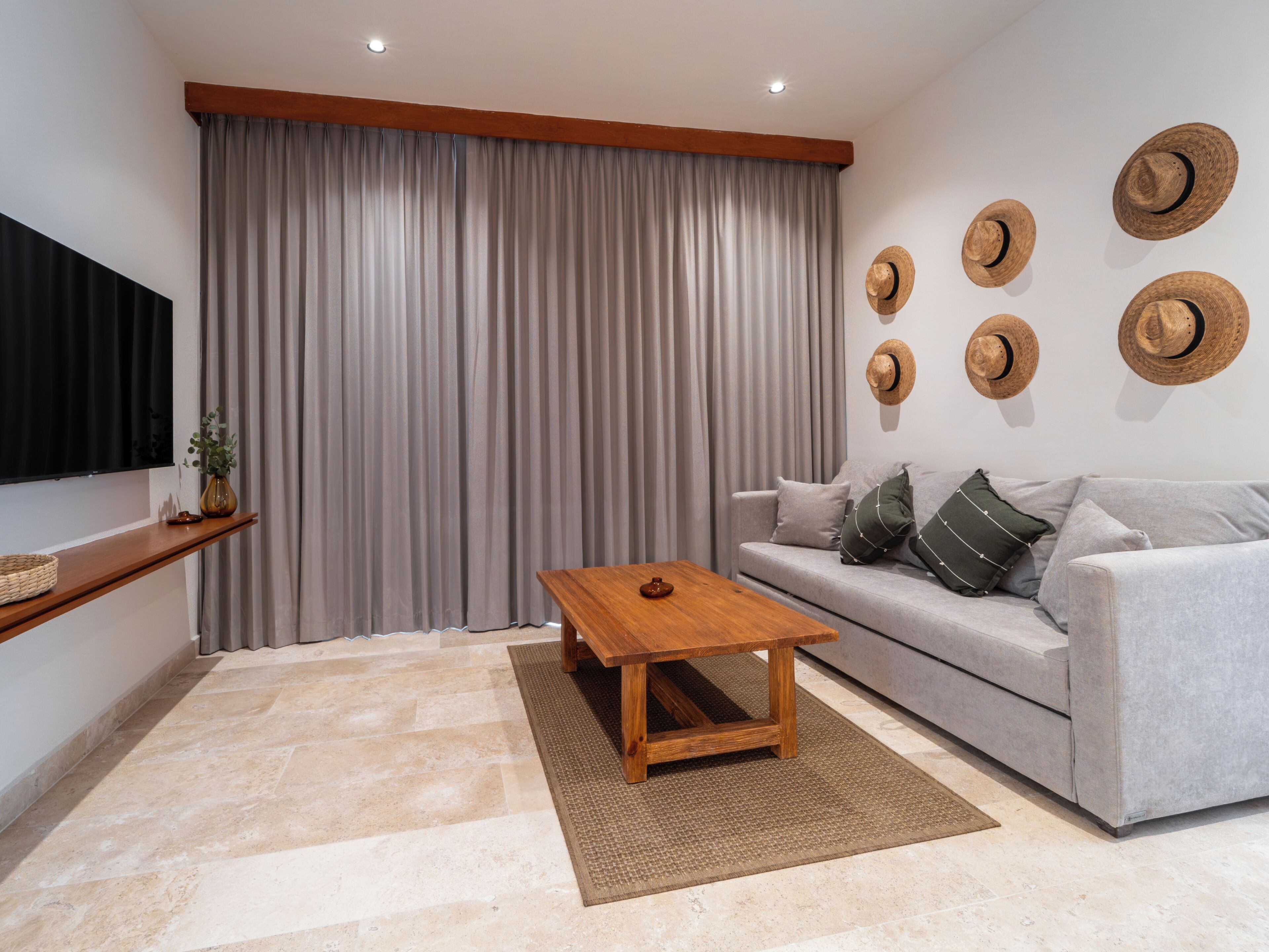 Un salon avec un canapé gris, une table basse en bois, un téléviseur fixé au mur et une grande fenêtre ornée de rideaux gris.