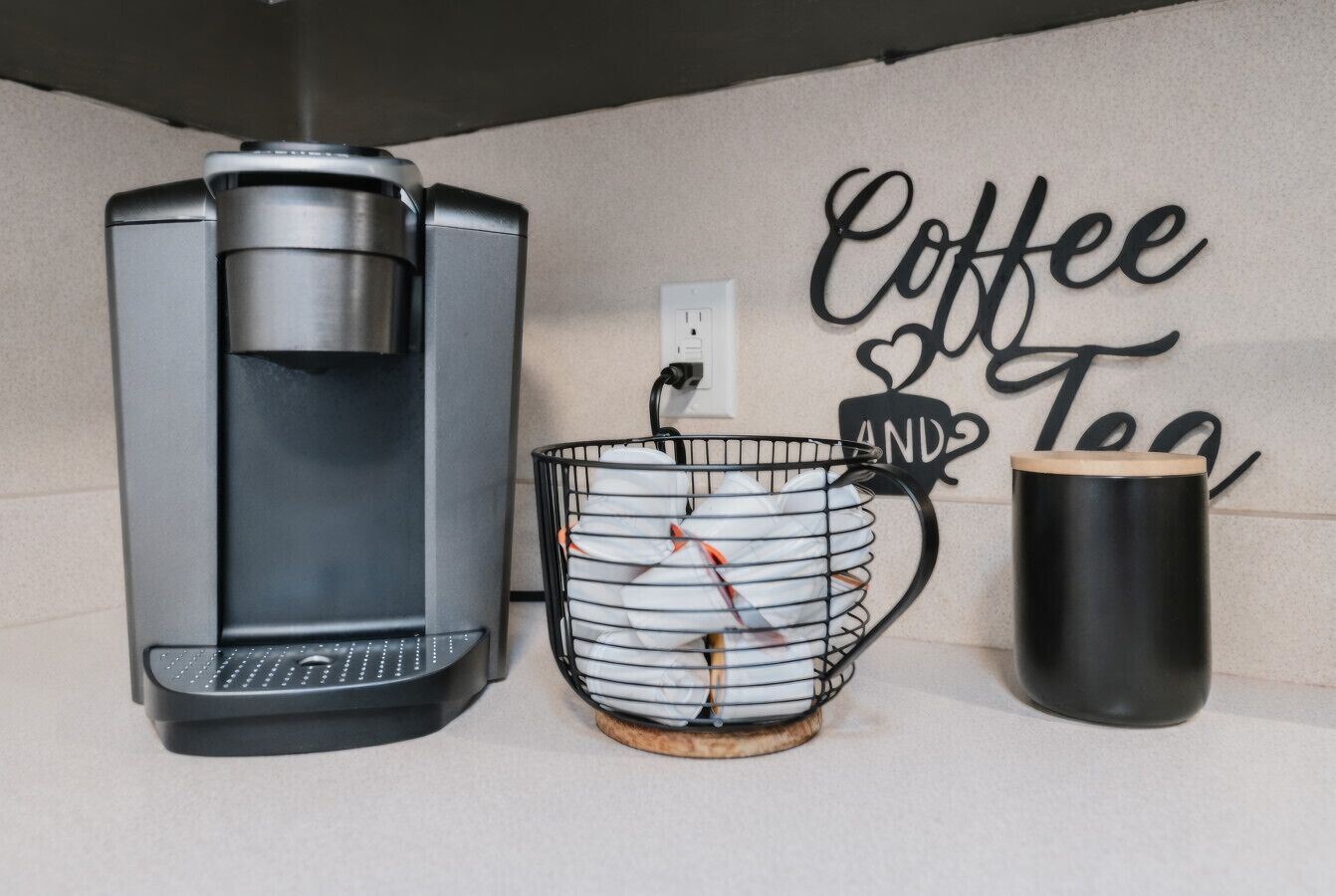 Eine Kaffeemaschine, ein Korb mit Kaffeekapseln und eine Tasse auf einer Arbeitsfläche. An der Wand hängt ein Schild mit der Aufschrift 'Coffee and Tea'.