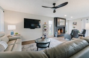 A modern living room with a fireplace, leather sofas, a flat-screen TV, and a ceiling fan.