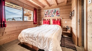 Ein Schlafzimmer in einer Holzhütte mit einem Bett, einem Fenster, einem roten Vorhang, einer an der Wand befestigten Ablage und einem Gemälde.