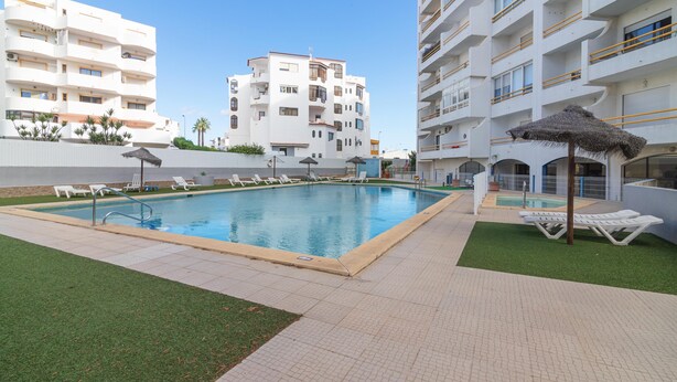 Un espace piscine avec des chaises longues et un parasol en chaume, entouré d’un bâtiment blanc doté de balcons.
