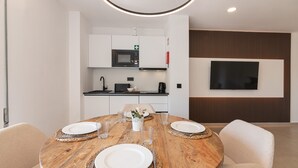 Un espace repas moderne avec une table en bois dressée pour quatre personnes, une cuisine en arrière-plan et un téléviseur à écran plat fixé au mur.