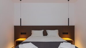 Une chambre d’hôtel moderne avec un lit aux couleurs sombres, deux oreillers blancs, une tête de lit en bois, des tables de chevet avec des lampes et des serviettes soigneusement pliées sur le lit.