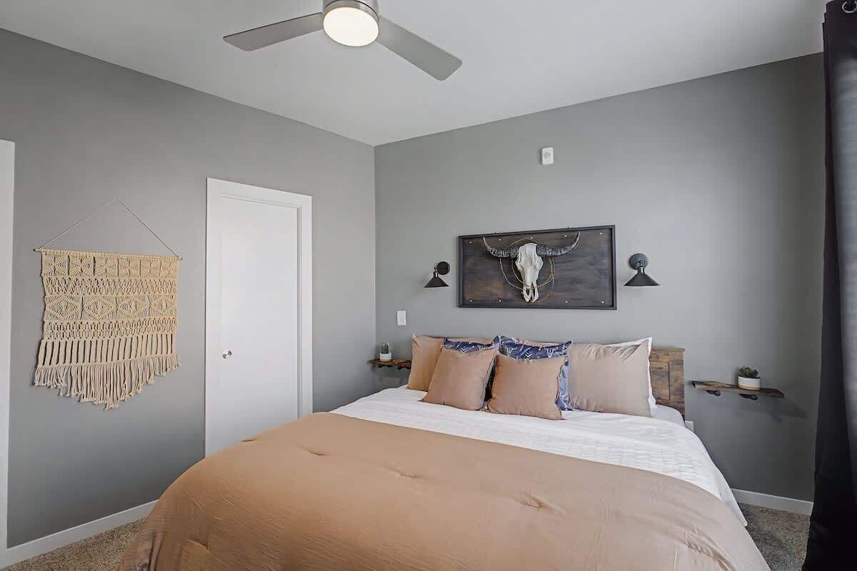 A bedroom with a bed, a ceiling fan, a macrame wall hanging, and a skull artwork on the wall.
