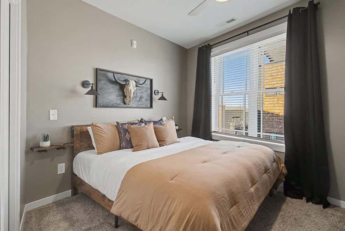 A bedroom with a large bed, a mounted artwork of a cow skull, a window with blinds, and a nightstand with a lamp.