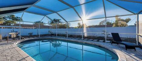 A swimming pool with a sunlit sky, a covered area with lounge chairs, and a clear view of the surrounding landscape.