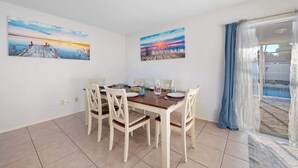 Un coin repas avec une table et des chaises en bois, deux tableaux accrochés aux murs et une vue sur une piscine par la fenêtre.