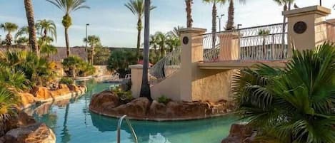 A swimming pool surrounded by palm trees and rocks, with a bridge and a clock tower in the background.