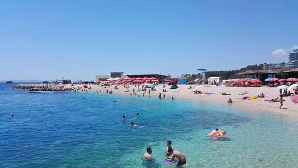 Uma praia com água azul cristalina, pessoas nadando e tomando sol, e guarda-sóis vermelhos.