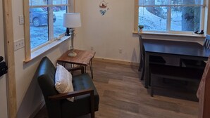 A room with a green chair, a small table with a lamp, a dining table with chairs, and a window with a view of parked cars and snow outside.