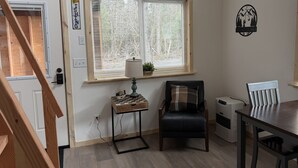 Een kamer met een houten trap, een raam met jaloezieën, een lamp op een bijzettafeltje, een stoel, een tafel en een decoratie aan de muur.