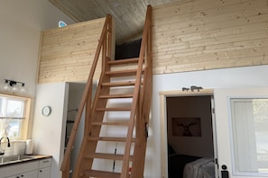 Une pièce avec une échelle en bois menant à une mezzanine, une fenêtre avec des stores, un évier et une horloge au mur.