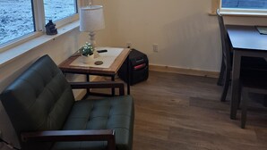 Ett rum med en grön soffa, ett litet bord med en lampa, en svart väska och ett fönster med utsikt över stranden.