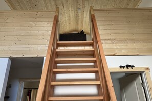 A wooden ladder leading to a lofted sleeping area with a black cushion, a wooden shelf with two small figurines, and a white door.