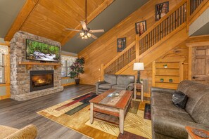 A spacious living room with a stone fireplace, a large flat-screen TV, a wooden ceiling fan, and a staircase with wooden railings. - Chateau Bliss – Elegant Mountain Retreat, Game room and Hot tub - Resort Pool (Gatlinburg)