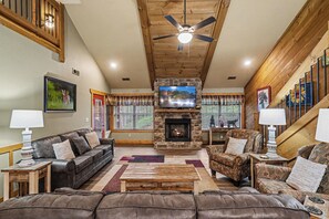 A living room with a stone fireplace, leather sofas, a television, and a ceiling fan. - Ivie Holler – Cozy 3-Bed Mountain Chalet with Game Room, Hot Tub, Resort Pool (Gatlinburg)