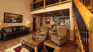 A spacious living room with a high ceiling, a wooden staircase, and a dining area with a kitchen in the background. - Misty Mountainside–Romantic 2-Suite Modern Chalet with Hot Tub and Pool Table (Gatlinburg)