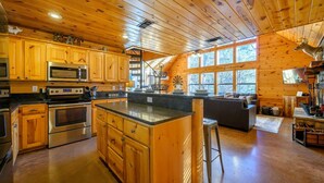 A spacious kitchen with wooden cabinets, a central island, and a large window providing natural light. - Bucky’s Blind · Cozy Forest Escape: Hot Tub, Playground, Fire Pit (Broken Bow)