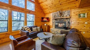 A cozy cabin interior with a stone fireplace, leather furniture, and large windows offering a view of the surrounding forest. - Bucky’s Blind · Cozy Forest Escape: Hot Tub, Playground, Fire Pit (Broken Bow)