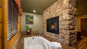 A bathroom with a stone wall, a fireplace, a large bathtub, and a window with blinds.